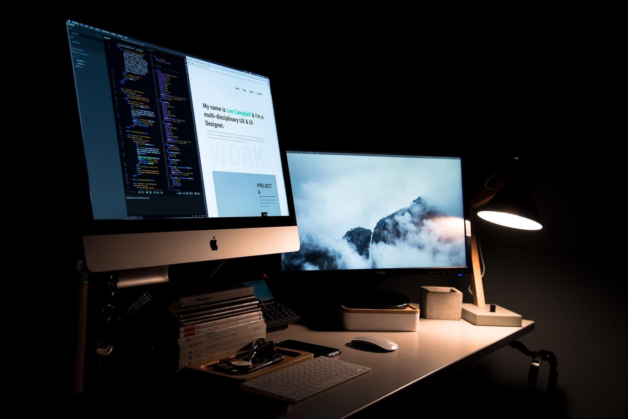 Dark desk with two monitors showing code and a design in a dimly lit workspace